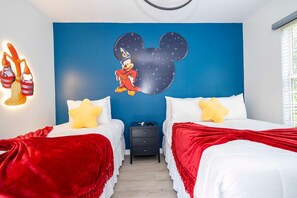 Une chambre avec deux lits, chacun recouvert d’une couverture rouge et orné d’un oreiller jaune. Un mur est décoré d’une fresque représentant Mickey Mouse, et une table de chevet avec un tiroir se trouve entre les deux lits.