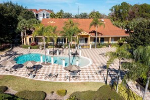 A pool area with checkered flooring, lounge chairs, umbrellas, and a building with a red roof in the background. - Gorgeous 4Bd w Pool at Encantada Resort 8523 (Kissimmee)