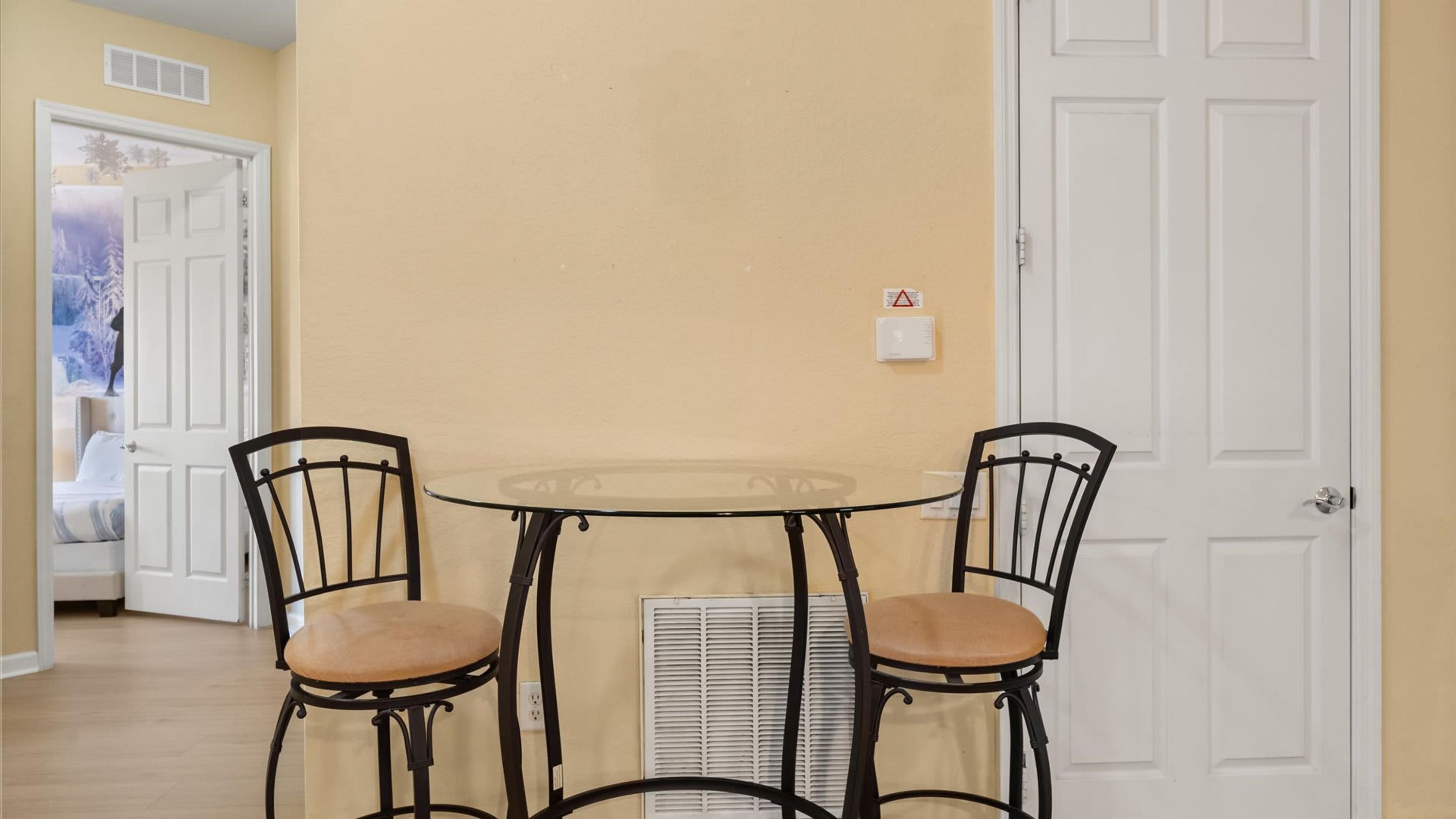 Een klein eetgedeelte met een tafel met glazen blad en twee stoelen, een witte deur en een ventilatie rooster aan de muur.