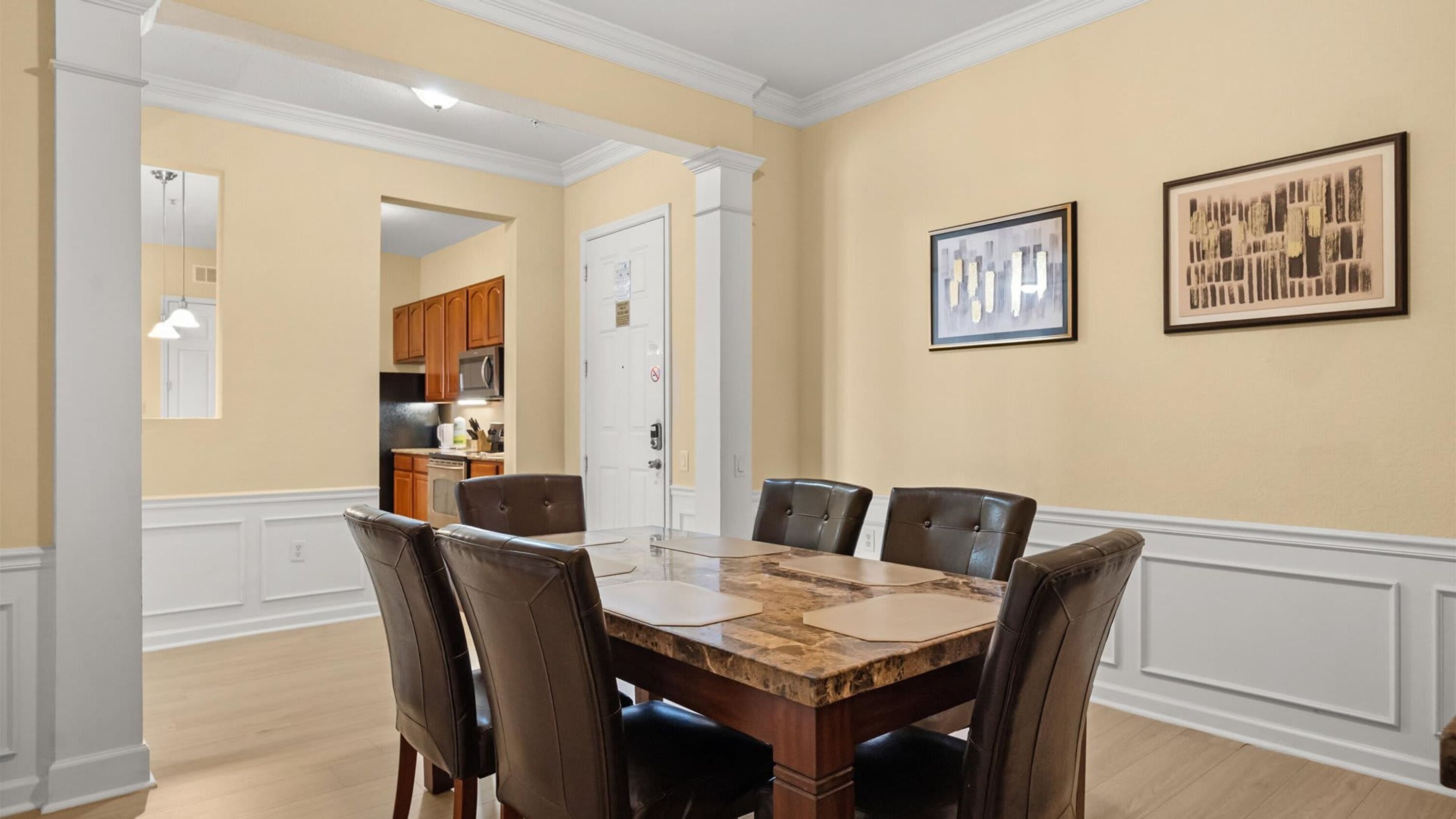 A dining area with a marble table and leather chairs, a kitchen in the background, and two framed artworks on the wall.