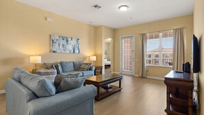 A living room with a sofa, a coffee table, and a TV. There is a painting on the wall and a window with blinds.