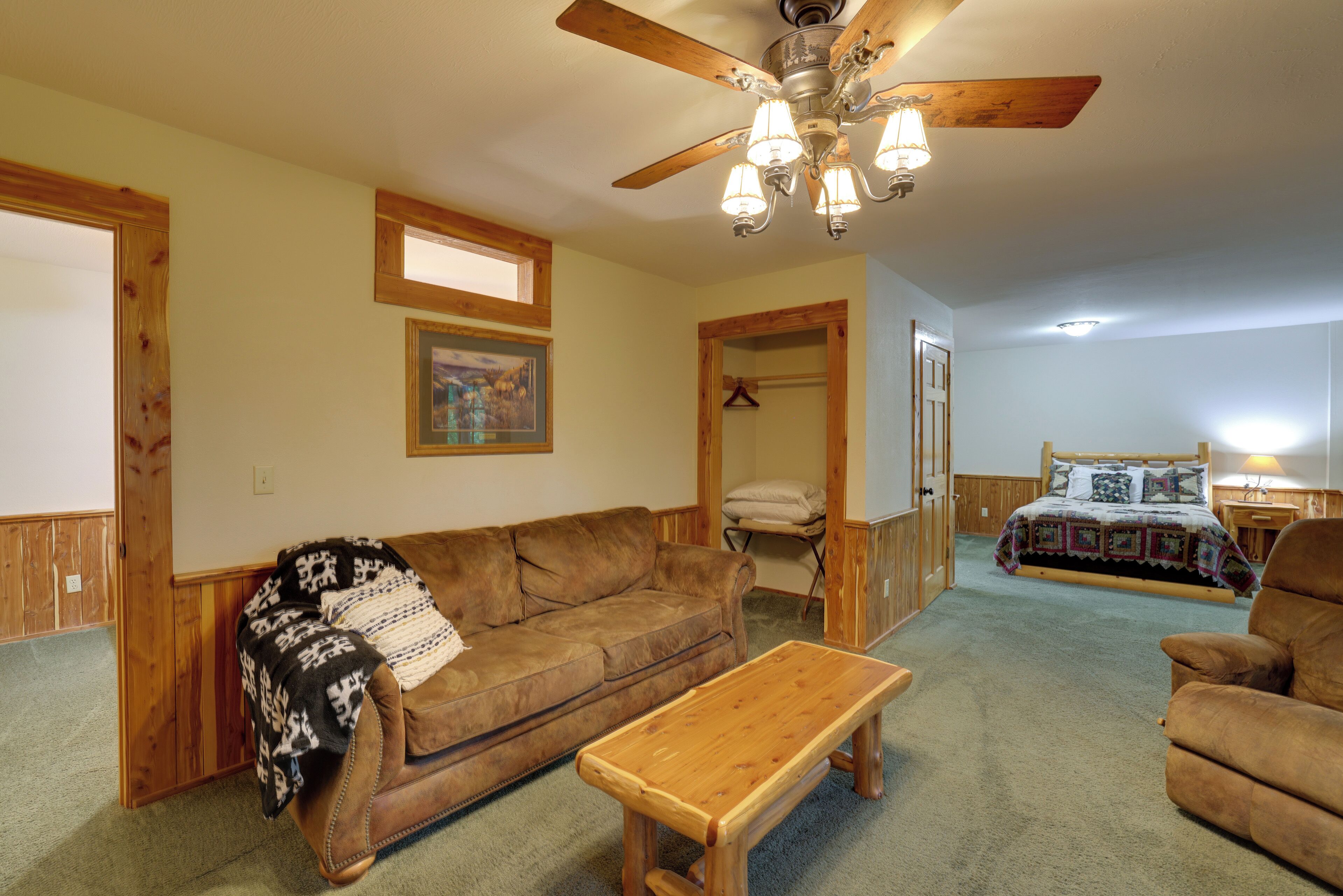 A cozy living room with a wooden ceiling fan, a brown leather sofa, a wooden coffee table, and a bed with a quilted blanket.