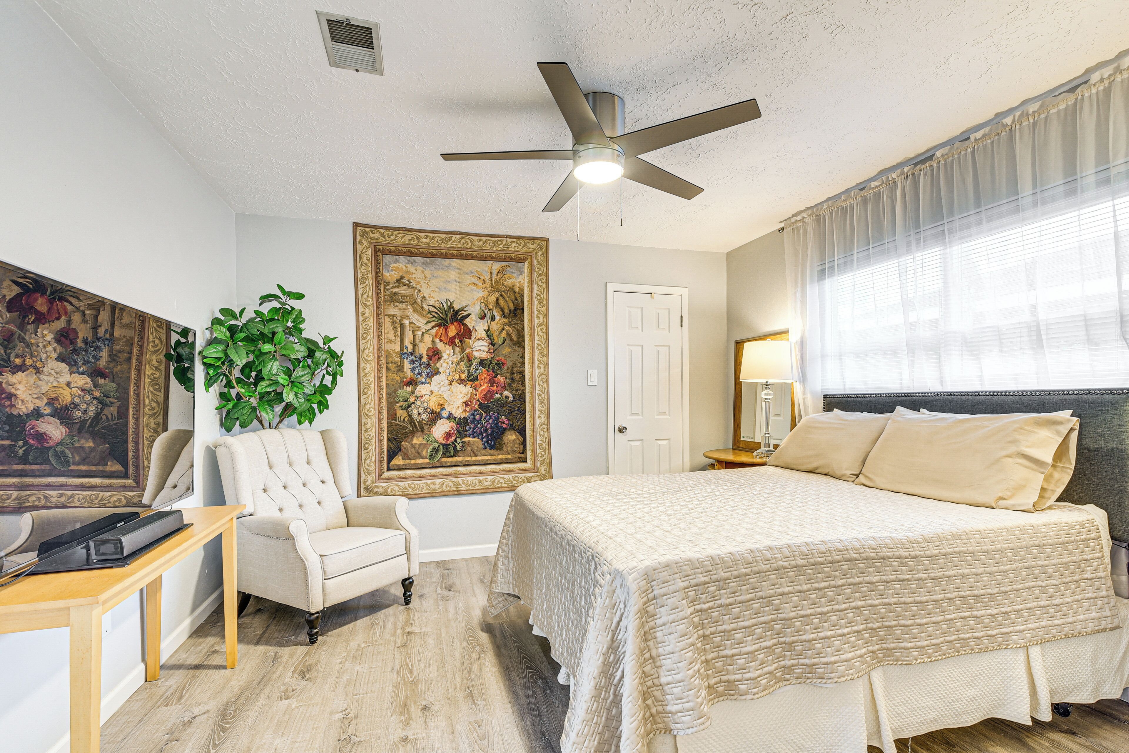 A bedroom with a bed, a ceiling fan, a large painting, a chair, and a desk.