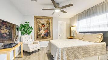 A bedroom with a bed, a ceiling fan, a large painting, a chair, and a desk.