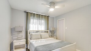 Un dormitorio con una cama, mesitas de noche, ventilador de techo y una ventana con cortinas.