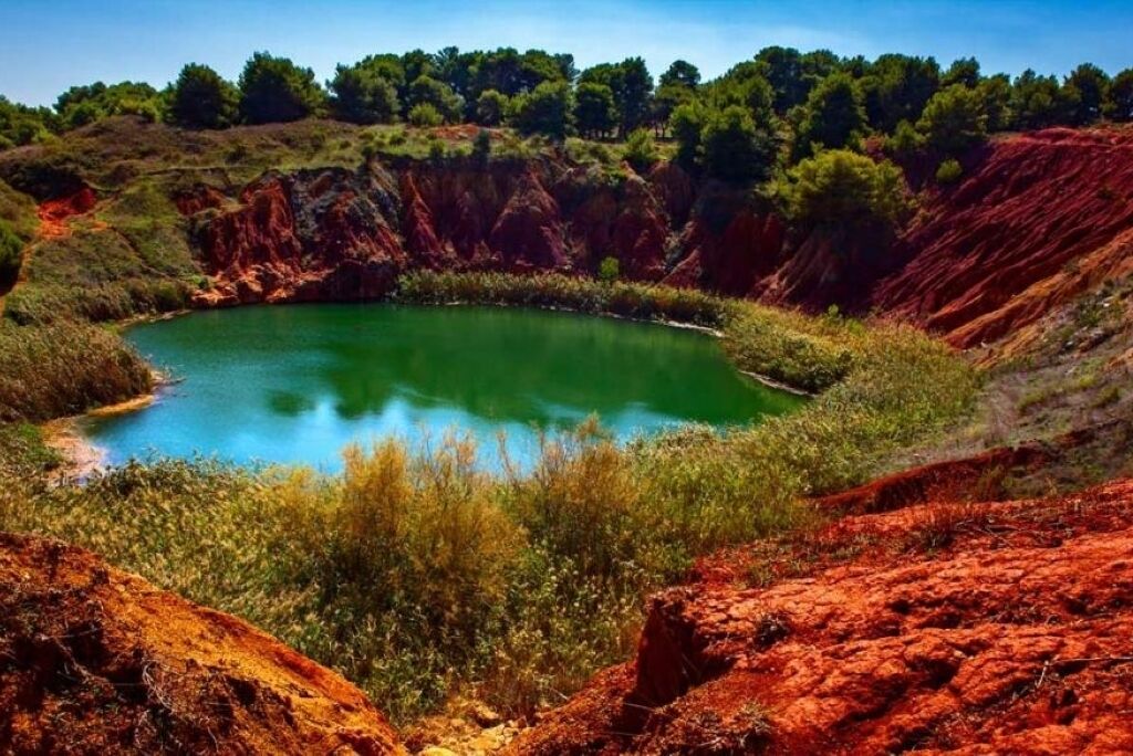 A natural landscape with a circular pond surrounded by reddish-brown cliffs and green vegetation.