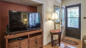 A living room with a flat-screen TV on a wooden stand, a small table with a lamp, and a door with a window view. - 05234 - 34 South Pego Way (Hot Springs)