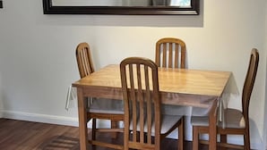 Un coin repas comprenant une table rectangulaire et quatre chaises en bois, un grand miroir au mur et une fenêtre avec des rideaux.