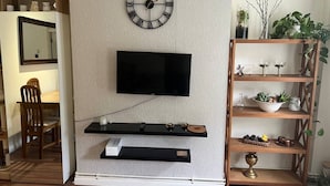 Un salon avec un téléviseur fixé au mur, des étagères en bois et une horloge au mur.