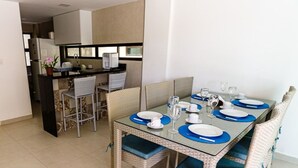 A dining area with a glass table set for four, surrounded by wicker chairs. A kitchen with a refrigerator and a toaster is visible in the background. - Casa BeiraMar 4QTS Piscina-SH031 (Ipojuca)