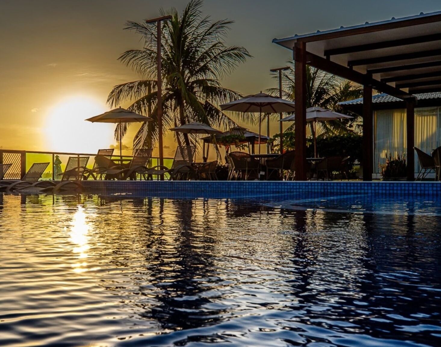 Zona junto a la piscina con sillones de descanso y sombrillas, palmeras y un edificio con piscina azulejada.