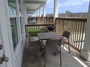 A covered outdoor patio with a table and four chairs, a grill, and a view of a grassy area beyond a wooden fence.