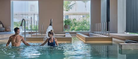 Piscina interior con agua cristalina, un hombre y una mujer nadando, y vistas a zonas verdes a través de amplios ventanales.