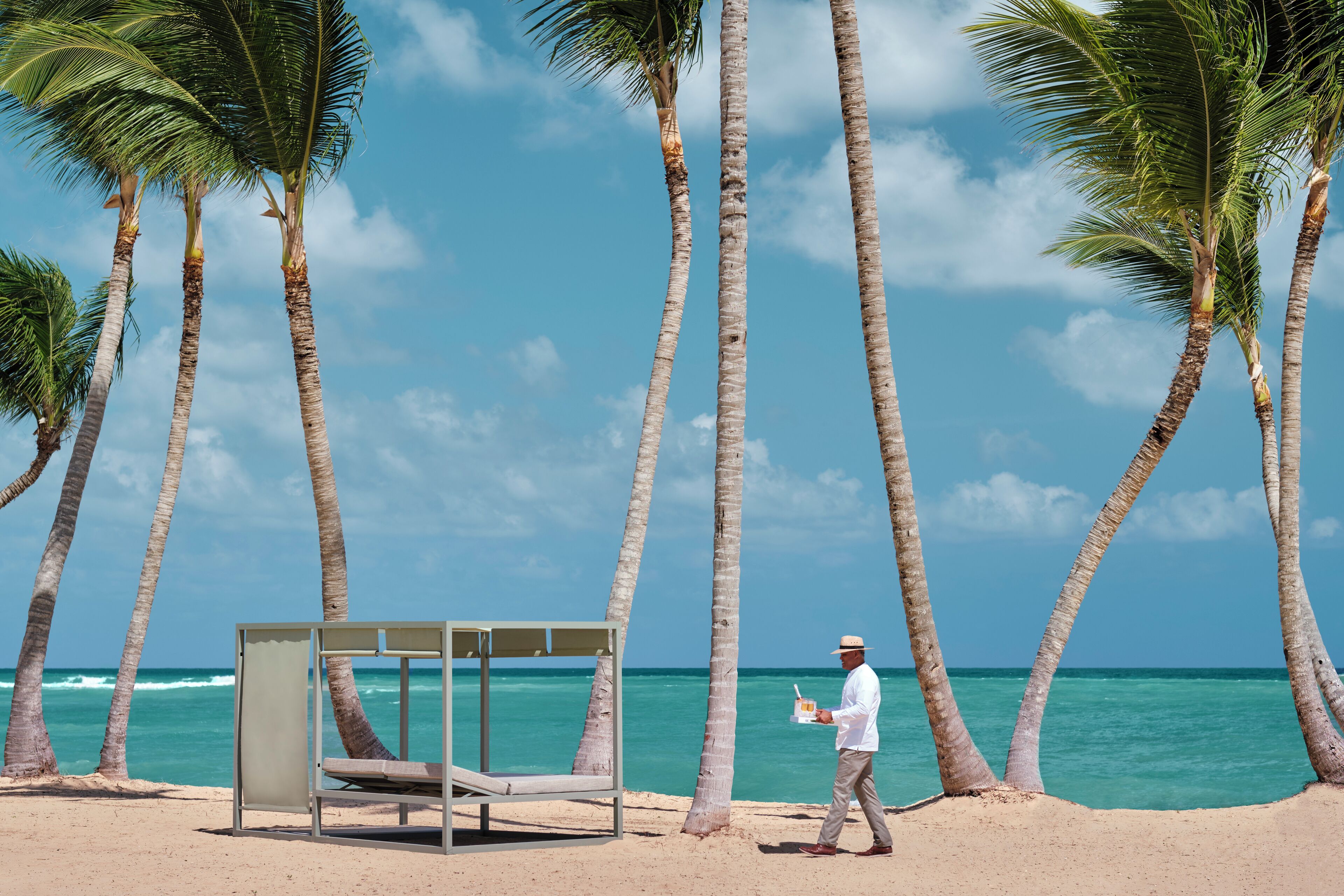 A beach with a single bed and a man holding a drink.