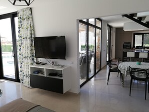 A modern living room with a flat-screen TV, a dining area with a round table and chairs, and a kitchen area with a countertop and cabinets.