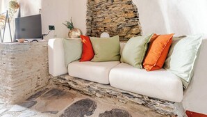 Un sofà blanc amb coixins verds i taronja, una paret de pedra i una televisió sobre un suport.