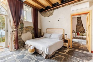 A bedroom with a stone wall, a bed with white linens, a wooden headboard, a nightstand, and a wicker basket.