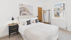 A bedroom with a large bed, a nightstand with drawers, a hanging light, a painting on the wall, and a window with a view of buildings. - Host & Stay | Admiral's View (Whitby)
