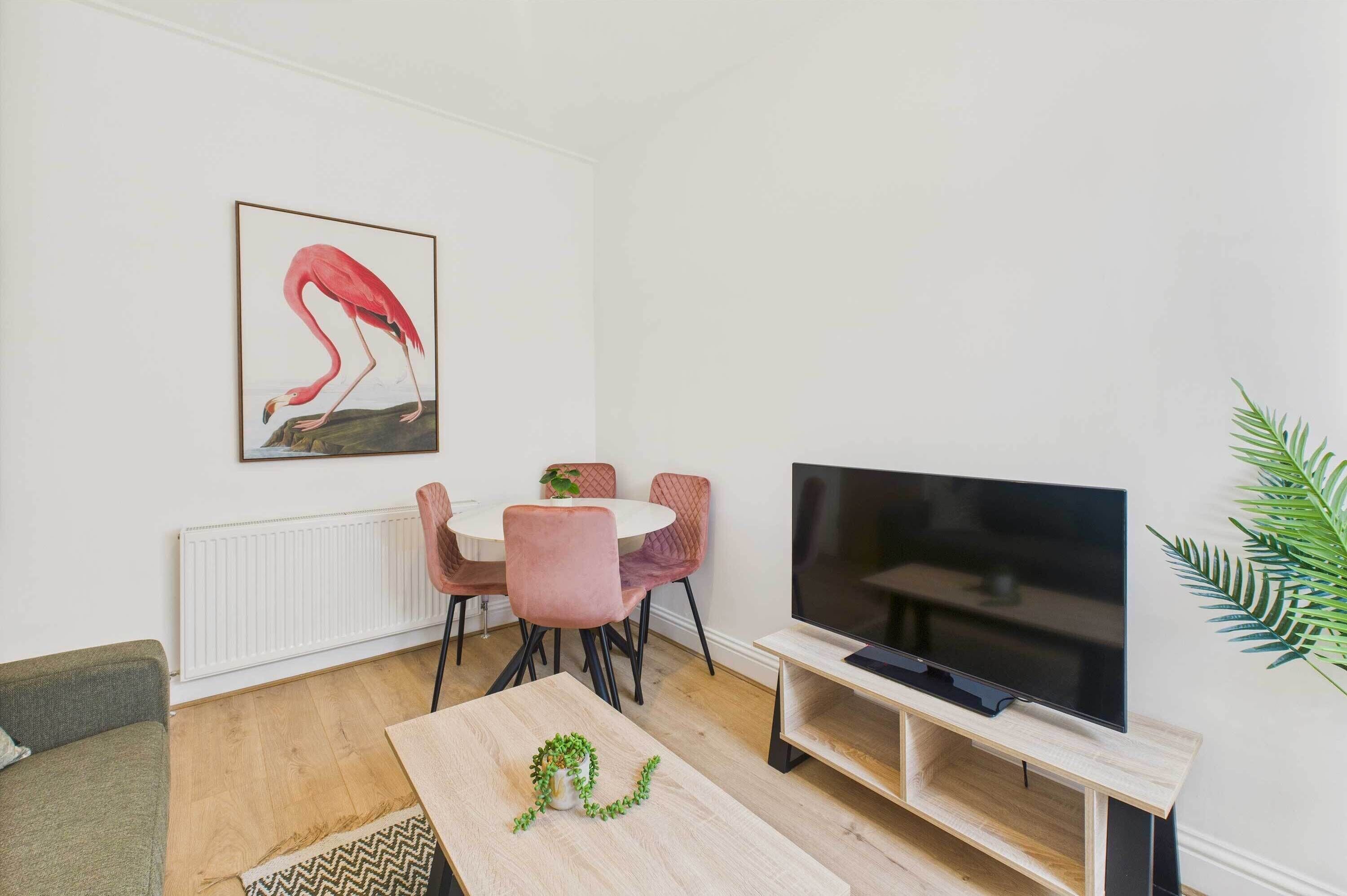 A living room with a white wall, a pink flamingo painting, a round table with pink chairs, a flat-screen TV on a wooden stand, and a green plant on a coffee table.