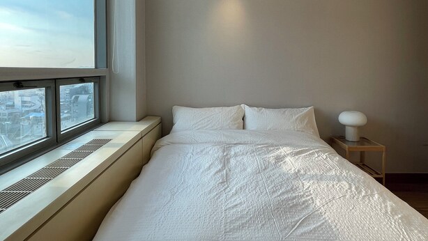 Habitación de hotel con un ventanal grande, una cama con ropa de cama blanca, una mesita de noche con lámpara y vistas a la ciudad.
