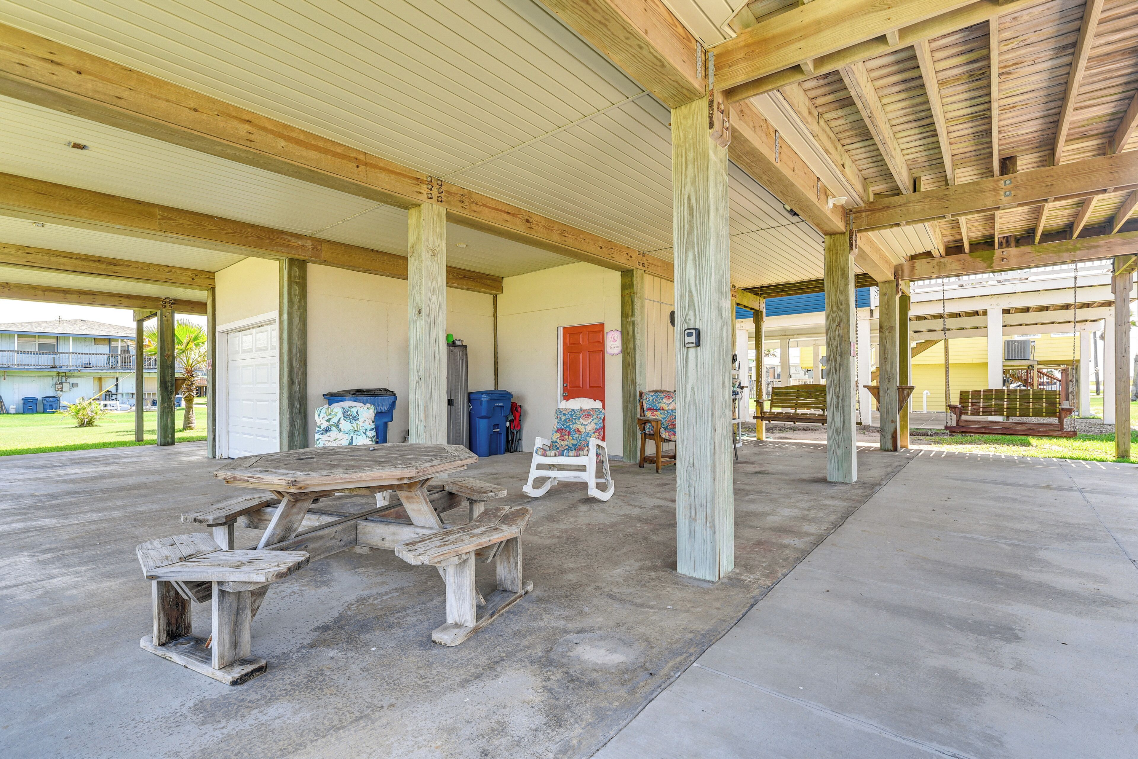 An outdoor covered patio area with a picnic table and benches, wooden pillars, and a view of a building in the background.