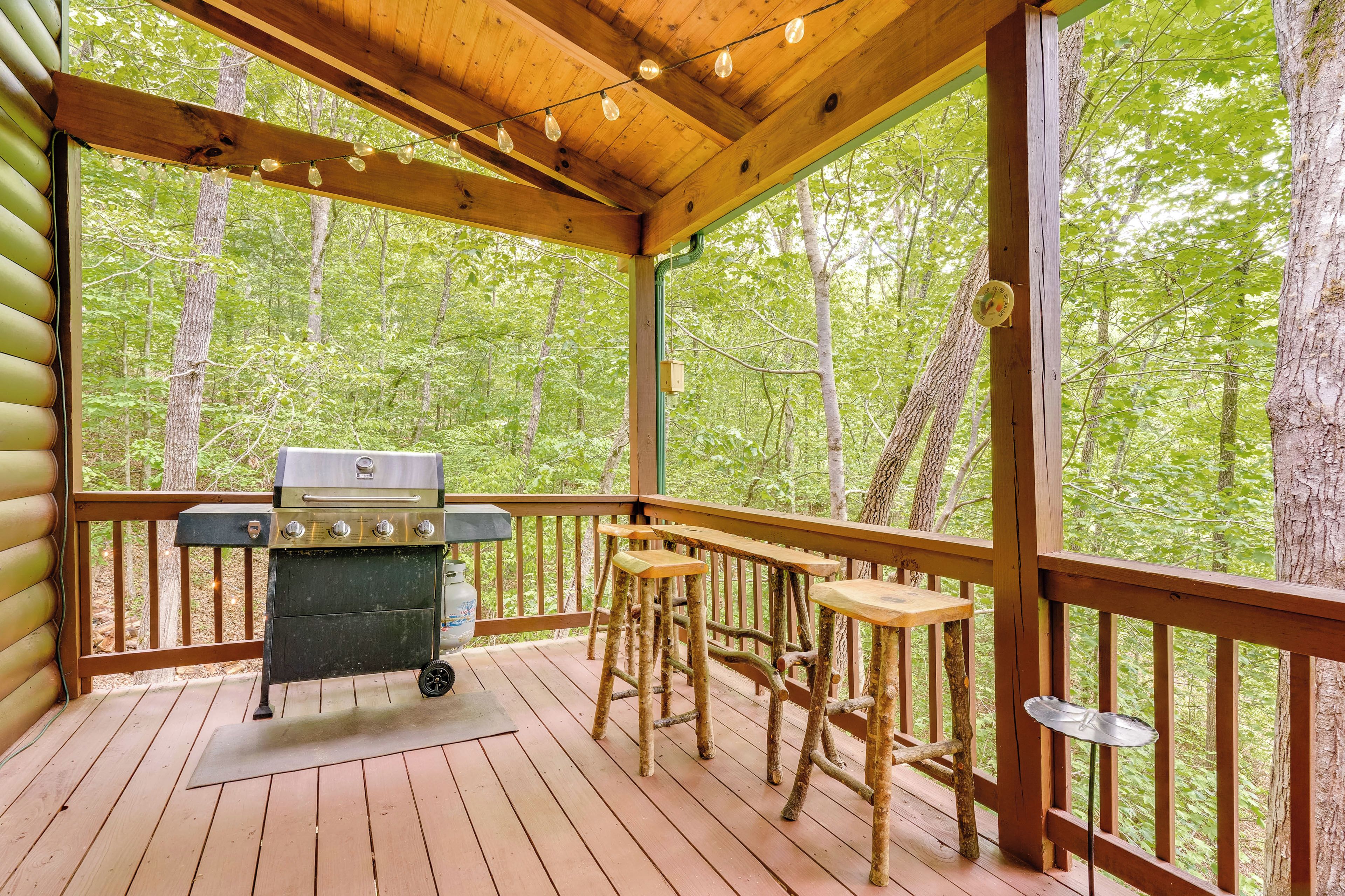 A covered deck with a grill, wooden stools, and a table, surrounded by trees.
