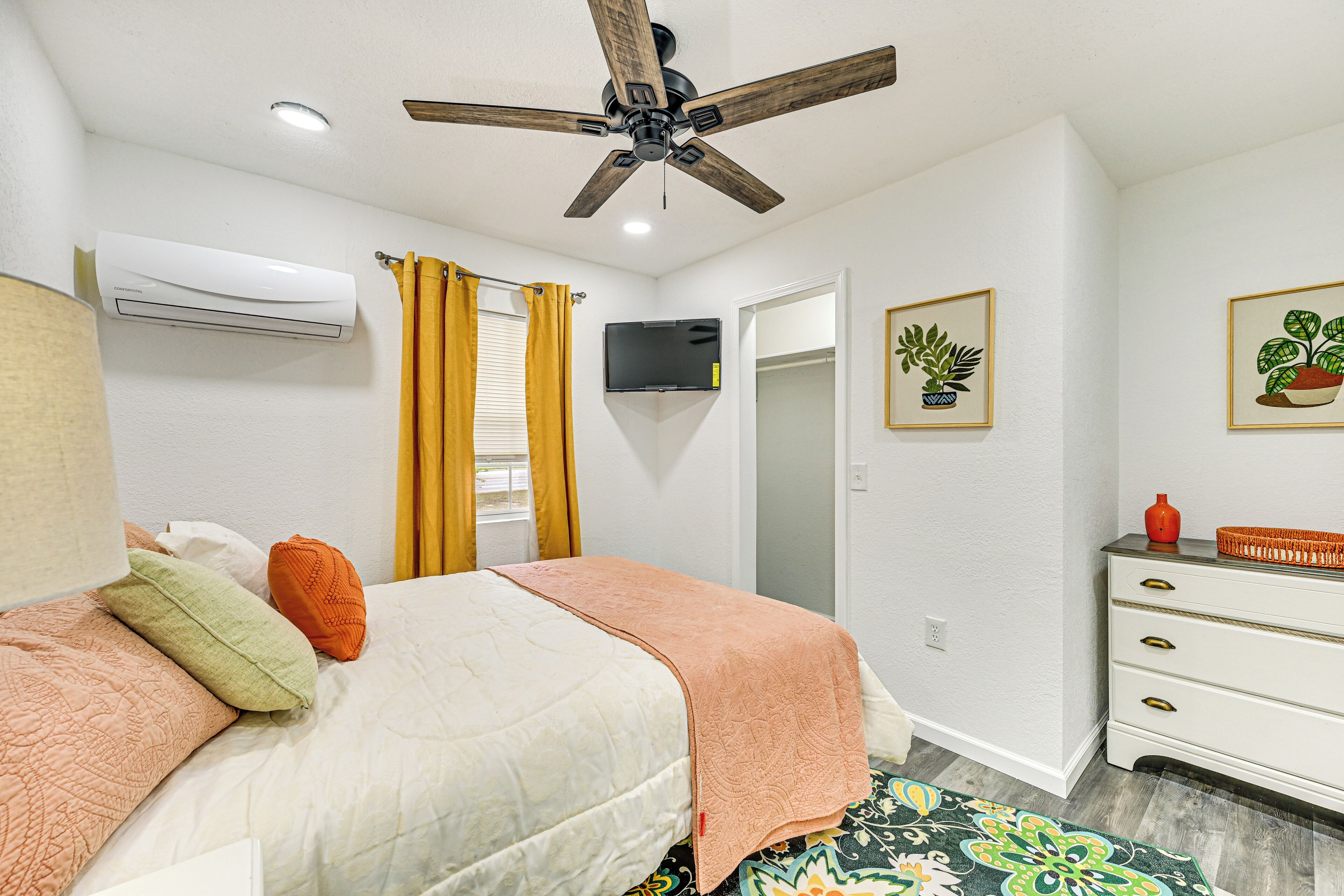 A bedroom with a bed, a ceiling fan, an air conditioning unit, a television, a dresser, and a door.