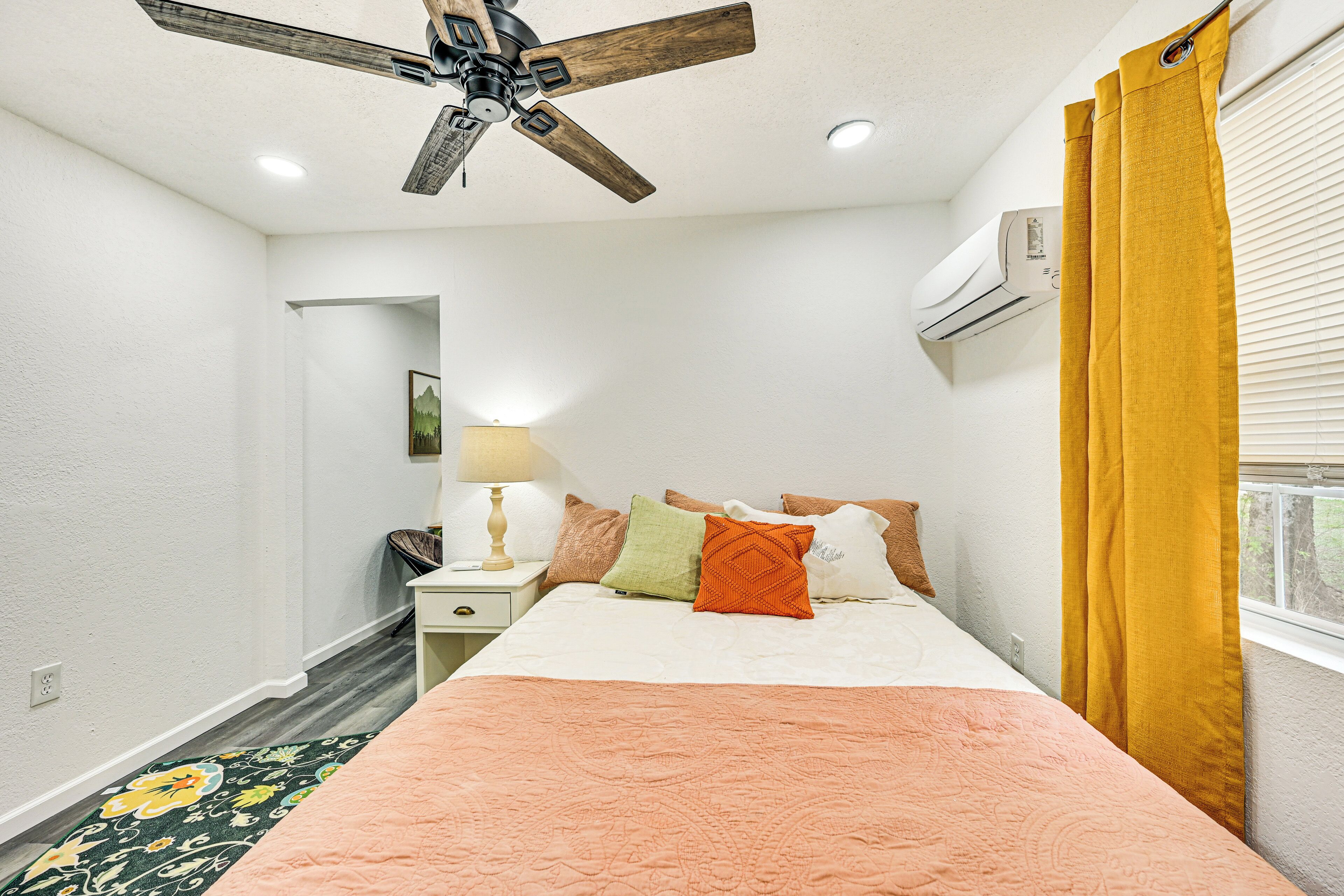 Een slaapkamer met een bed, een plafondventilator, een airconditioner, een raam met gele gordijnen en een nachtkastje met een lamp.