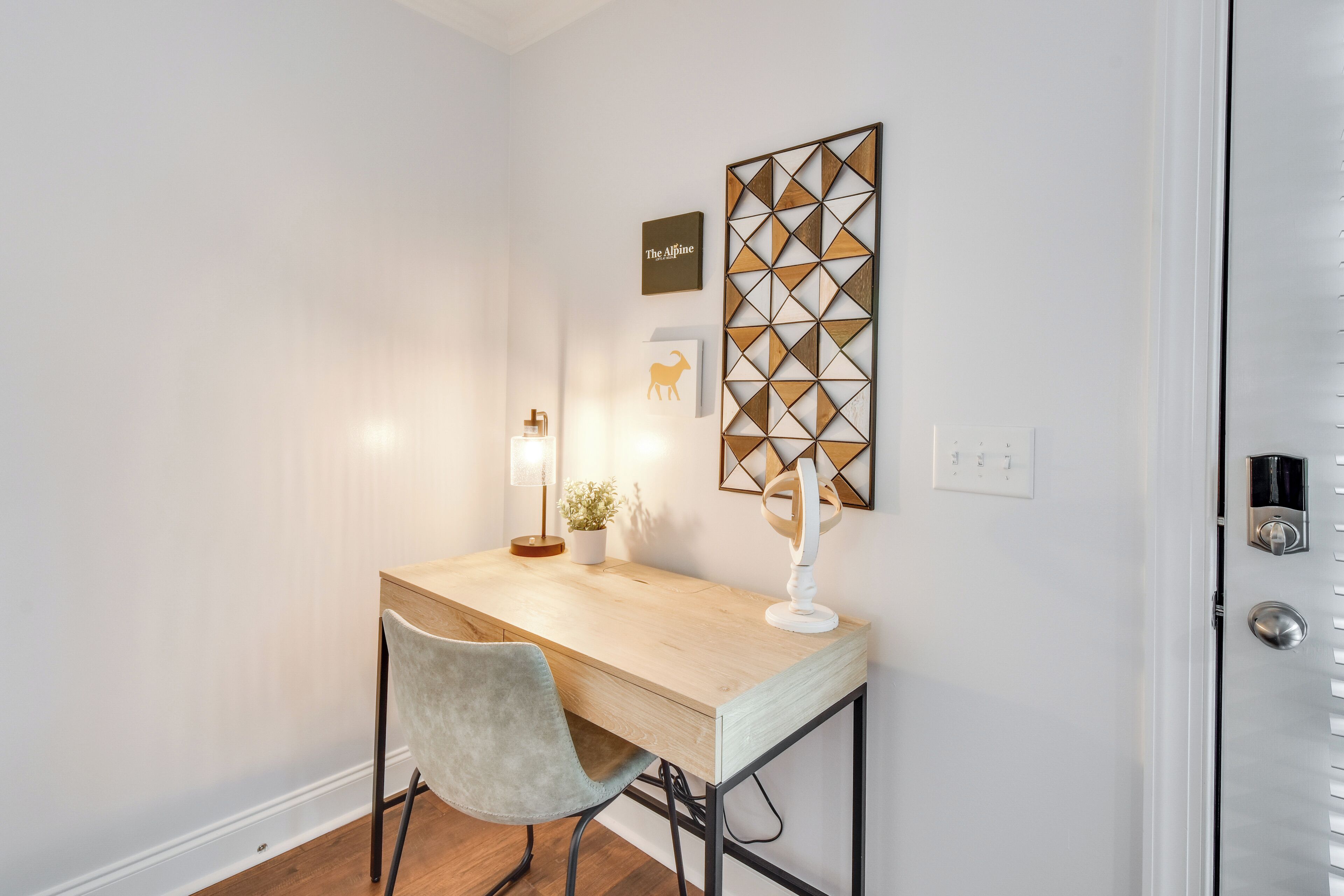 Un espacio de trabajo pequeño y ordenado, con un escritorio de madera, una silla blanca y una lámpara fijada a la pared.