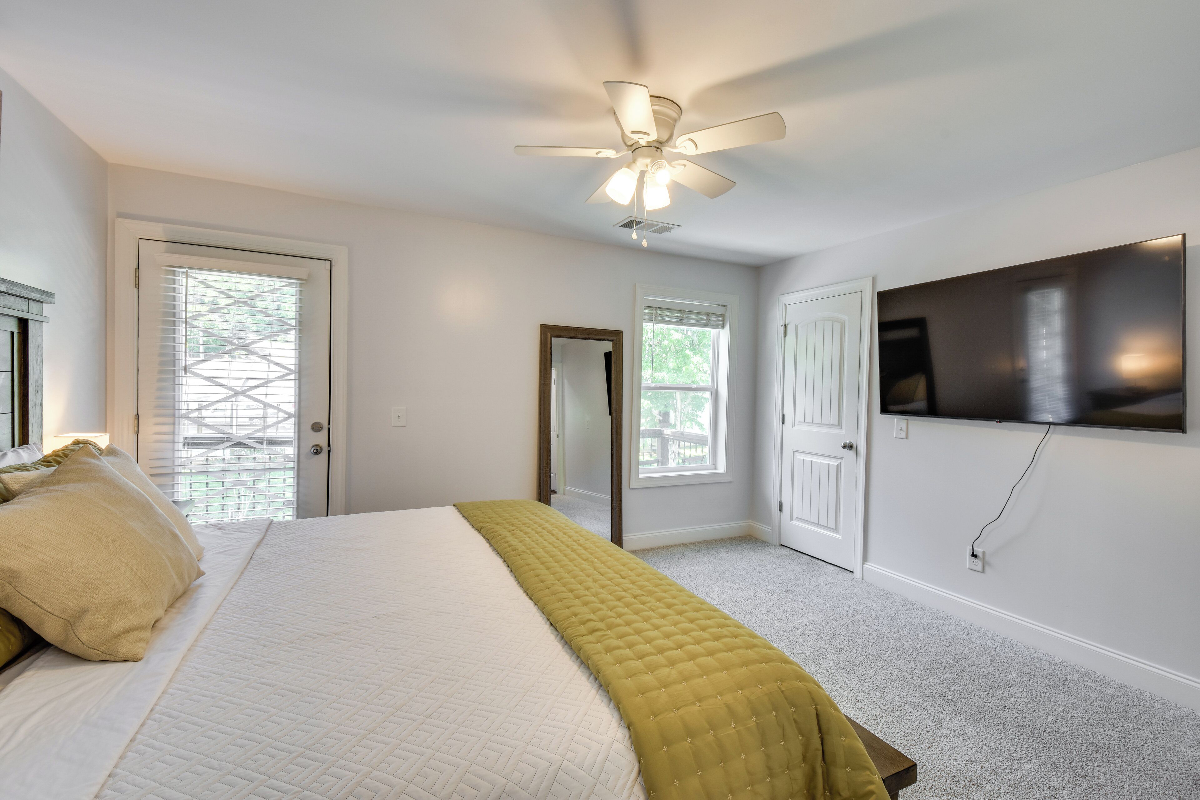 A bedroom with a bed, a ceiling fan, a TV mounted on the wall, and a door leading to another room.