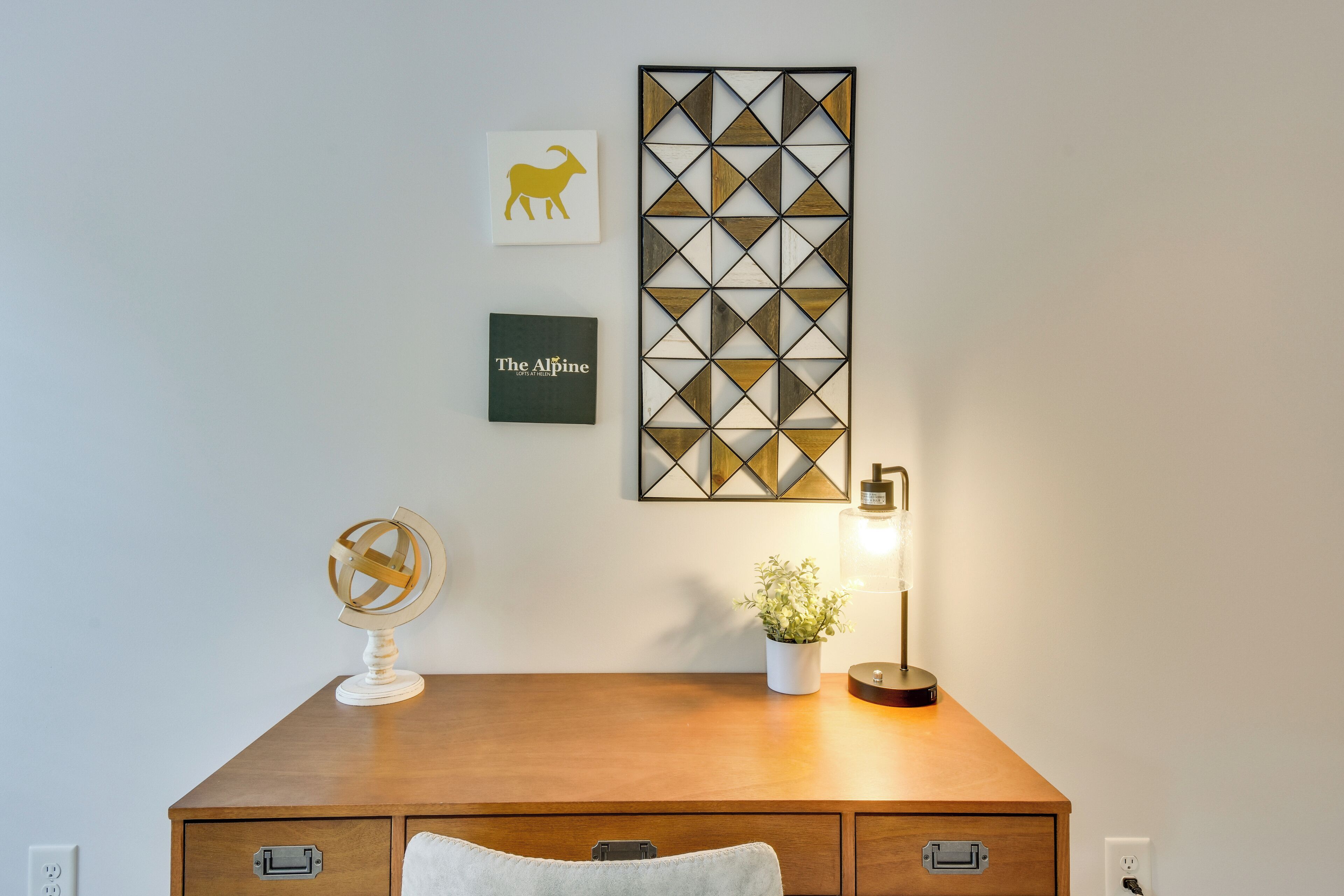 A wooden desk with a globe, a framed picture of a goat, a framed text 'The Alpine', a potted plant, and a lamp.