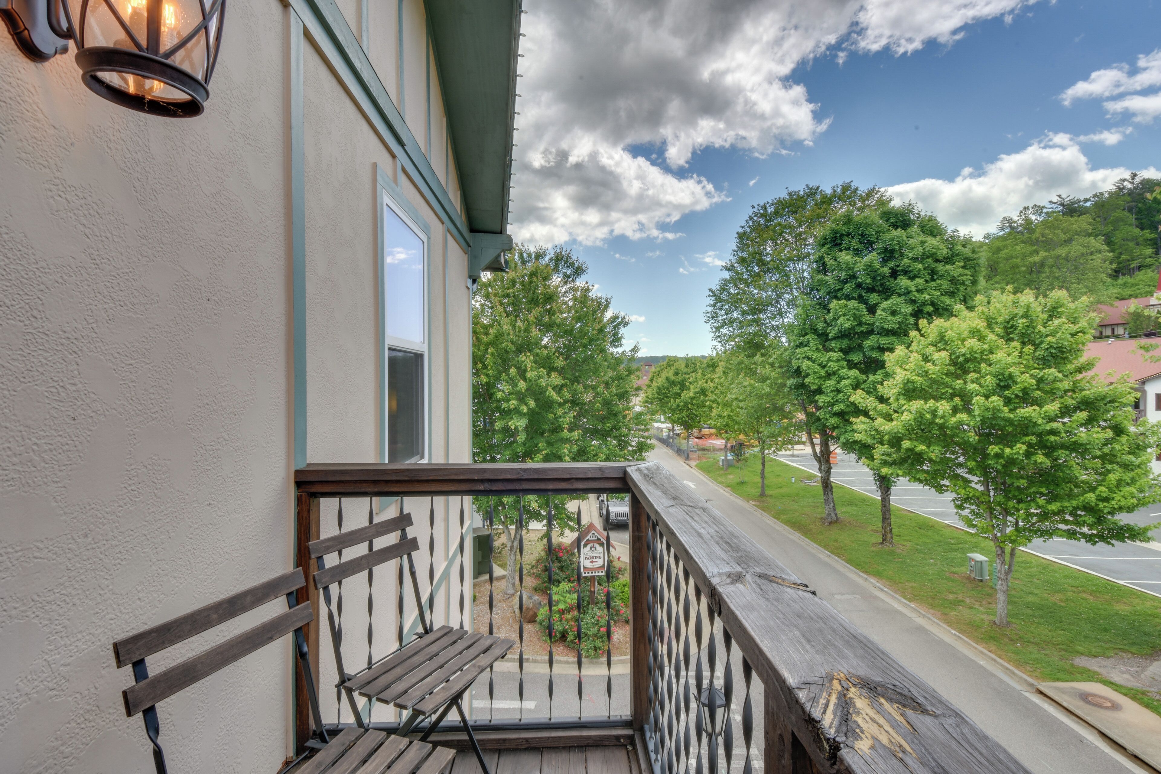 A wooden bench on a balcony with a metal railing, overlooking a street with trees and a clear sky.