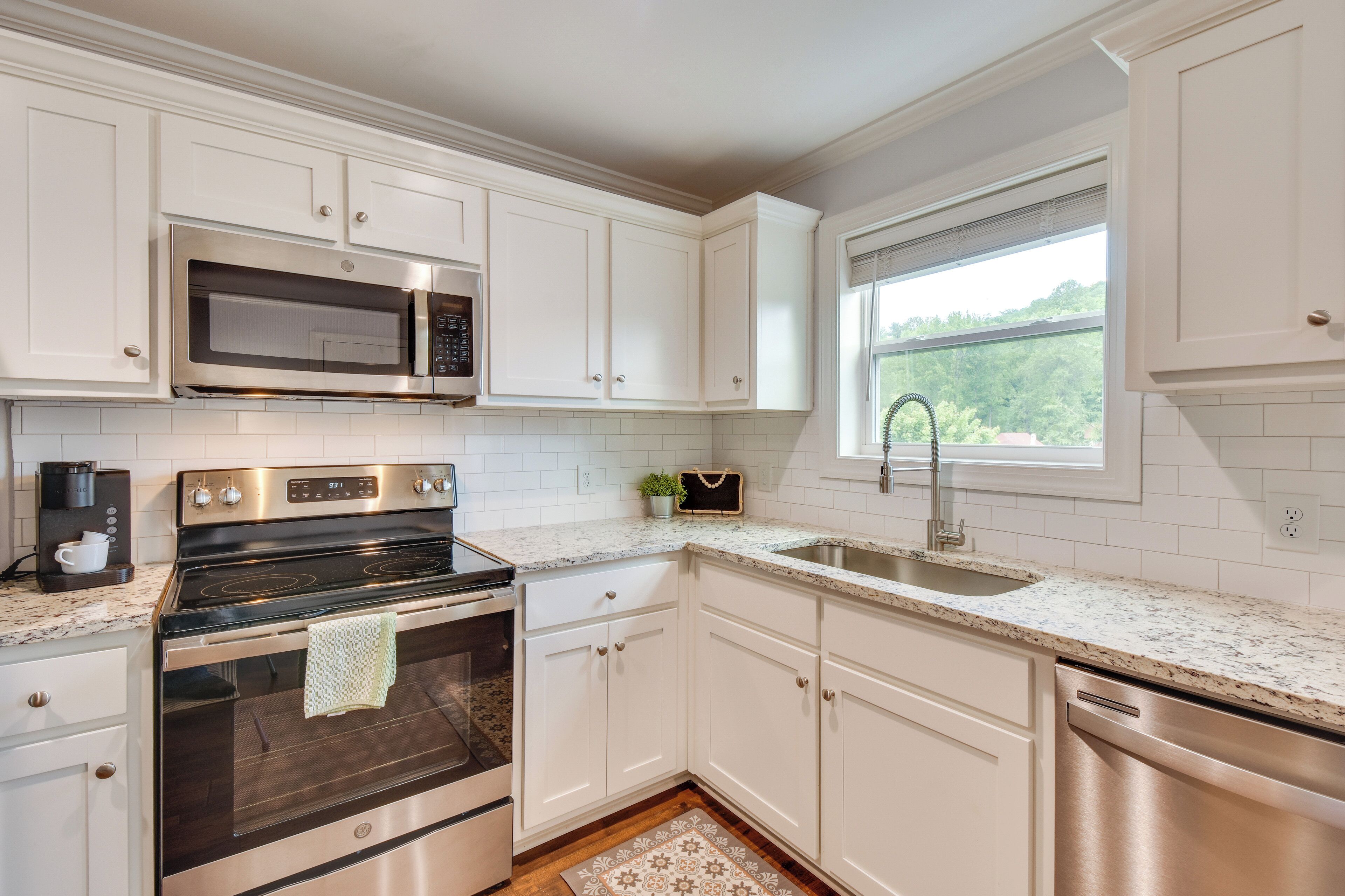 A modern kitchen with white cabinets, stainless steel appliances, a granite countertop, and a window with a view of greenery.