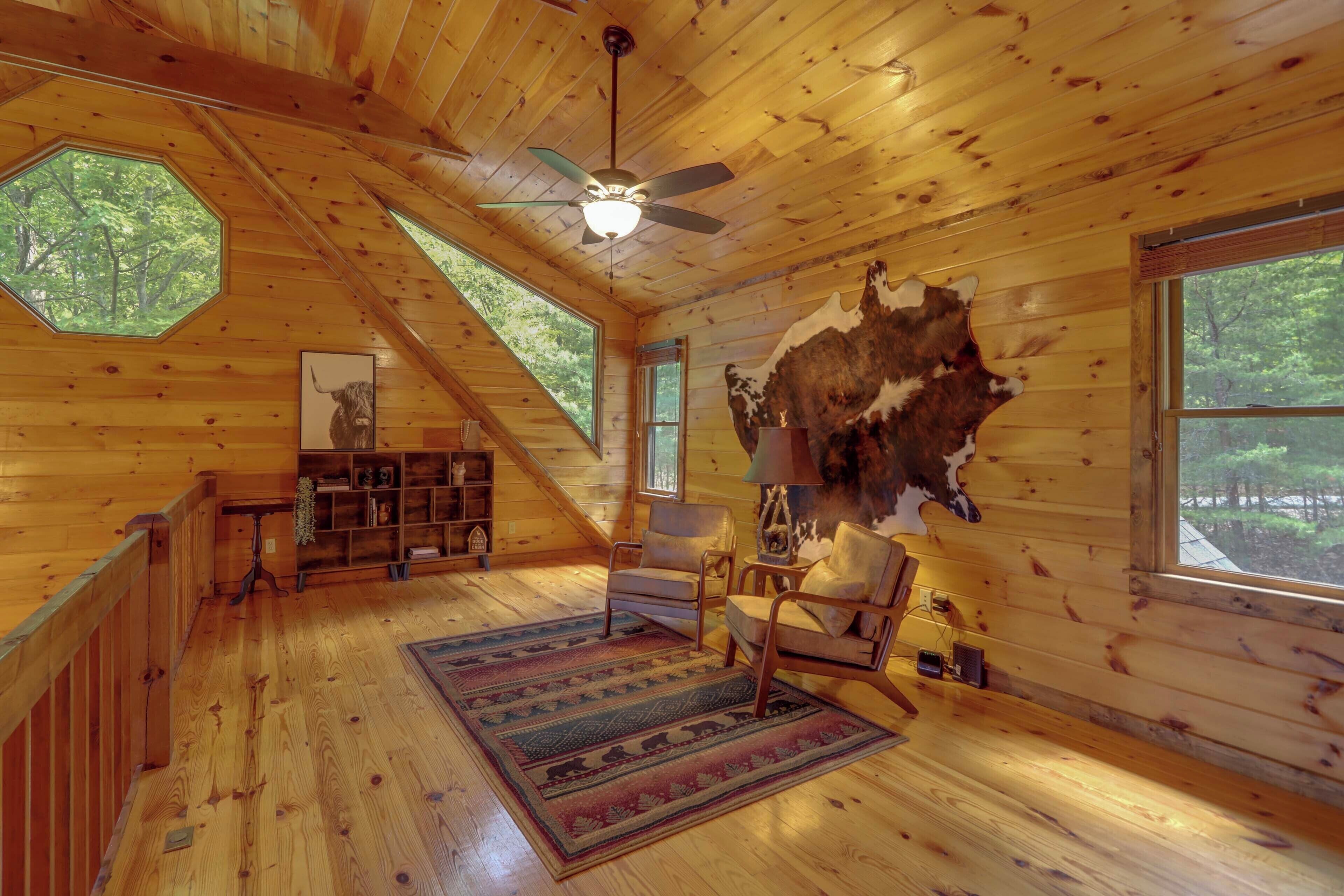 L’intérieur d’une cabane en rondins, avec un plancher en bois, un ventilateur de plafond, une grande fenêtre et une peau d’animal fixée au mur.