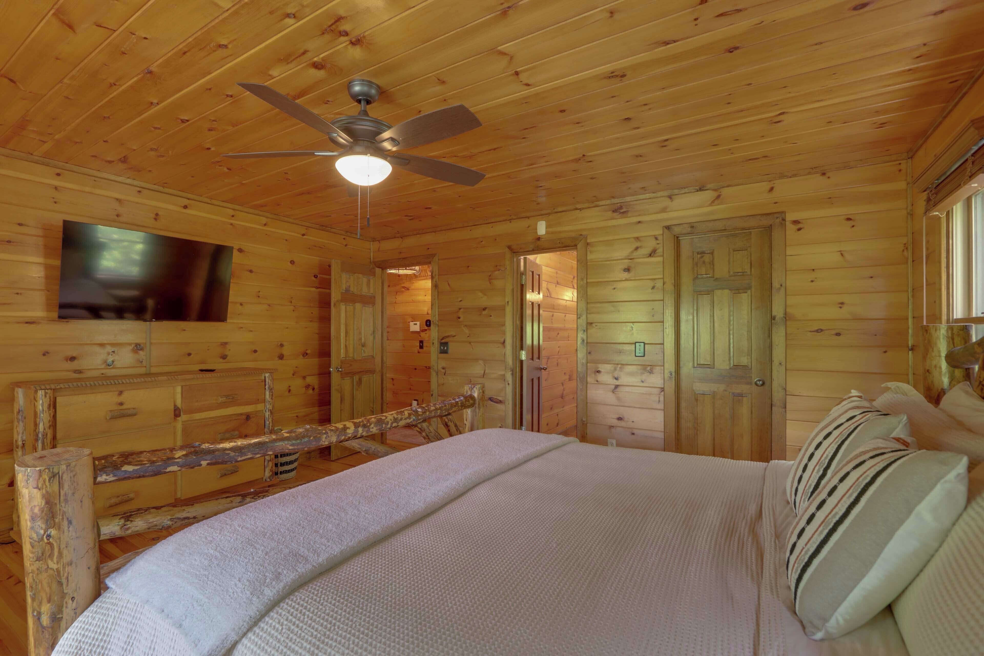 A wooden cabin bedroom with a bed, a ceiling fan, and a television mounted on the wall.