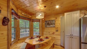 A wooden cabin interior with a dining table, benches, a refrigerator, and a large window. - Family Friendly: Hot Tub Haven in Mineral Bluff! (Mineral Bluff)