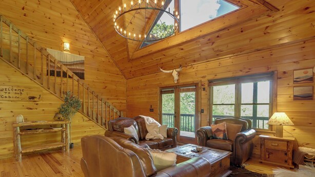 A cabin interior with wooden walls, a leather sofa, a chandelier, and a staircase. - Family Friendly: Hot Tub Haven in Mineral Bluff! (Mineral Bluff)