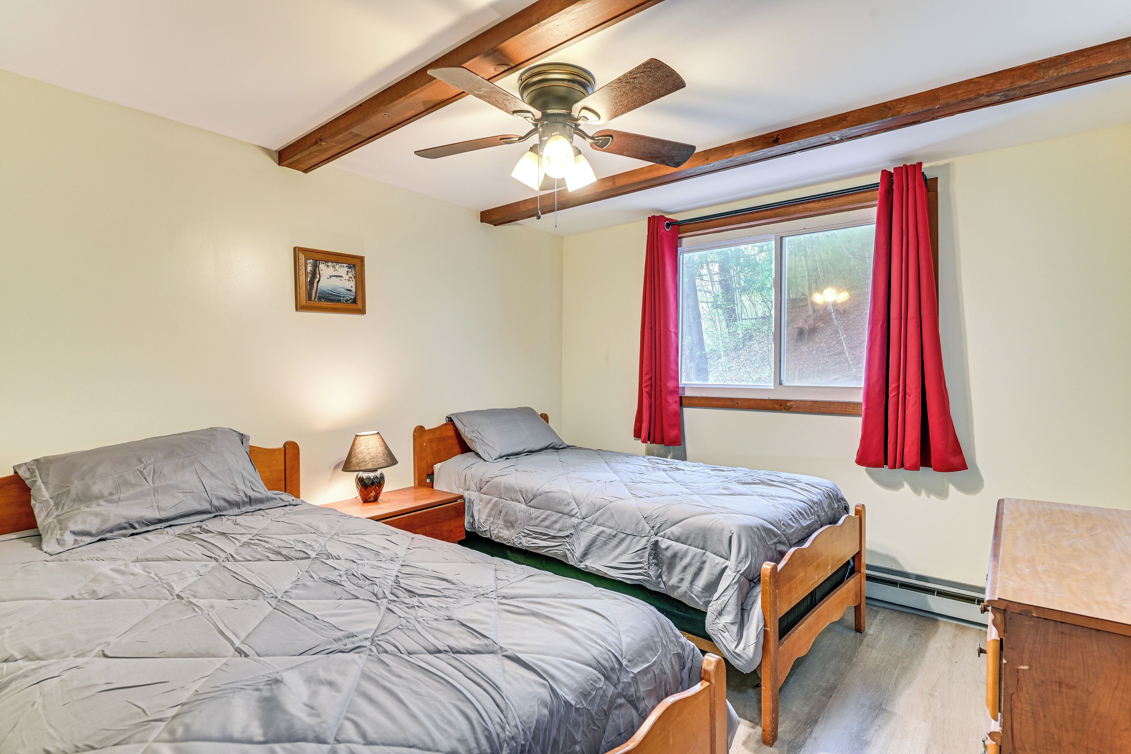 A bedroom with two single beds, a ceiling fan, a window with red curtains, and a small bedside table with a lamp.