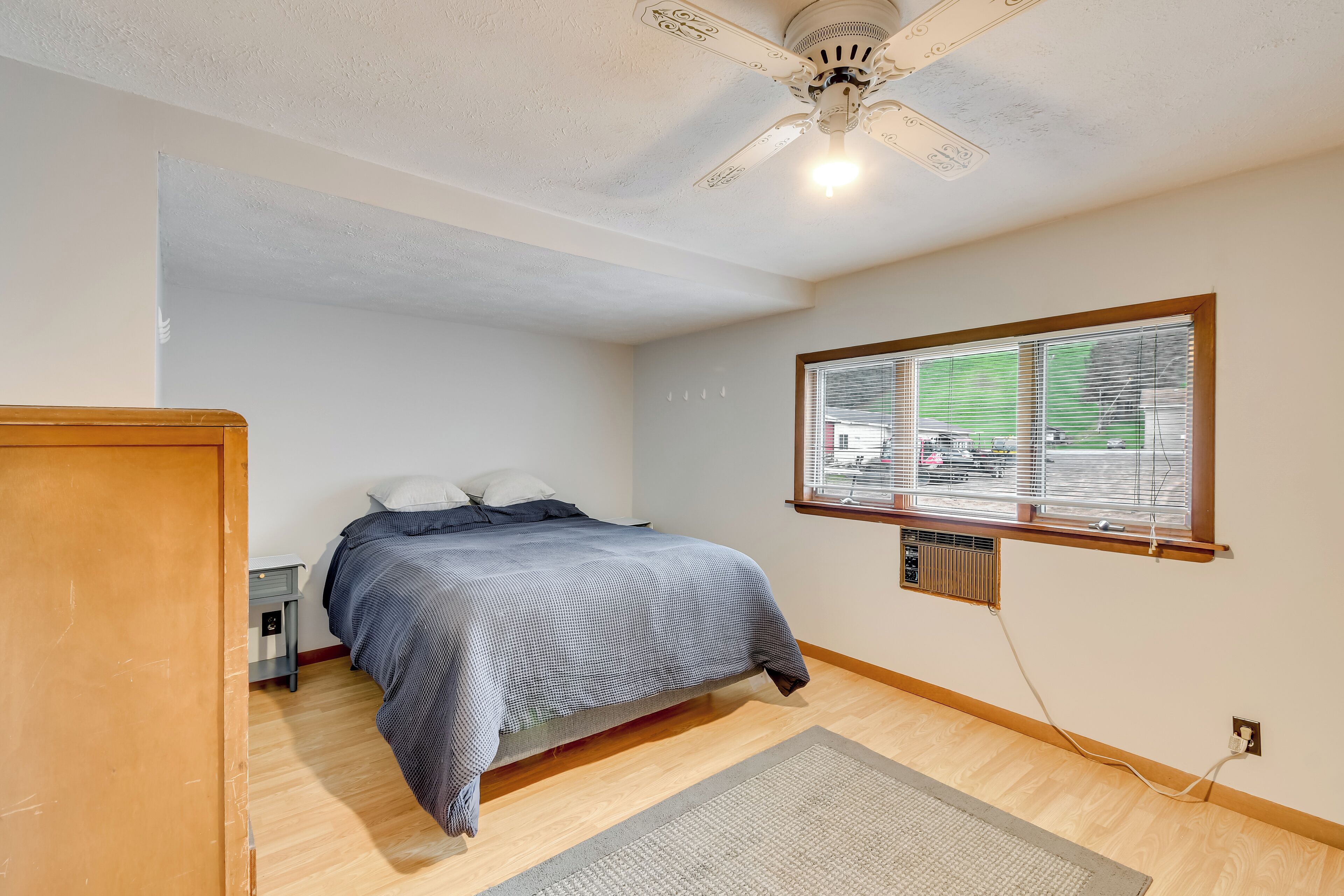 Une chambre à coucher comprenant un lit, une table de chevet, un ventilateur de plafond, une fenêtre avec des stores et un radiateur.