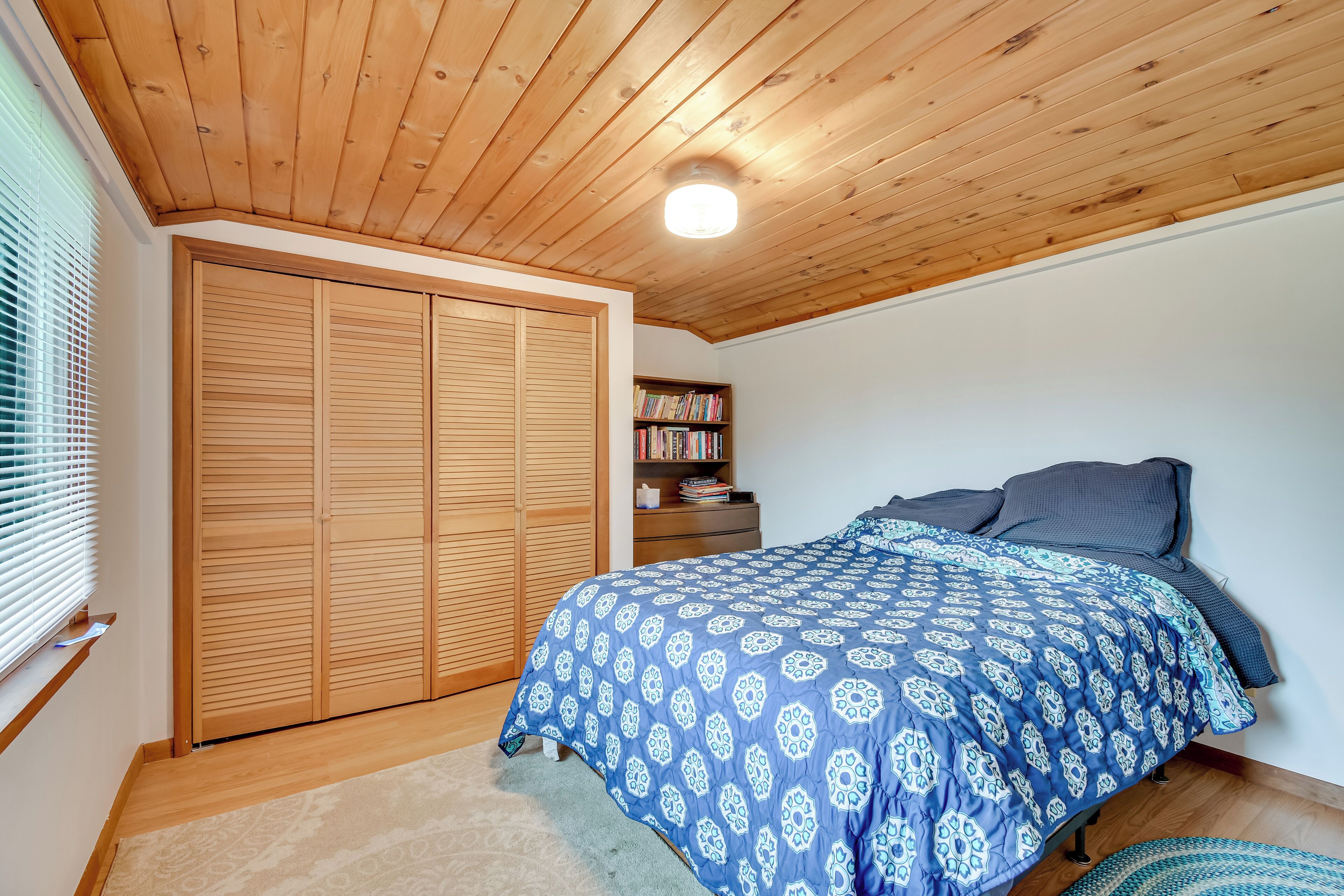 Un dormitorio con techo de madera, una cama grande con edredón azul y blanco estampado, una estantería y un armario de madera.