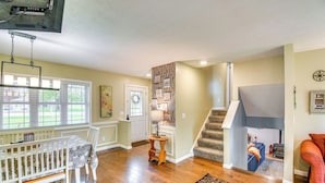 A spacious living room with hardwood flooring, a white crib, a dining table with chairs, and a staircase leading to an upper level. - 3 Mi to Erie Canal: Home w/ Hot Tub in Holley! (Holley)