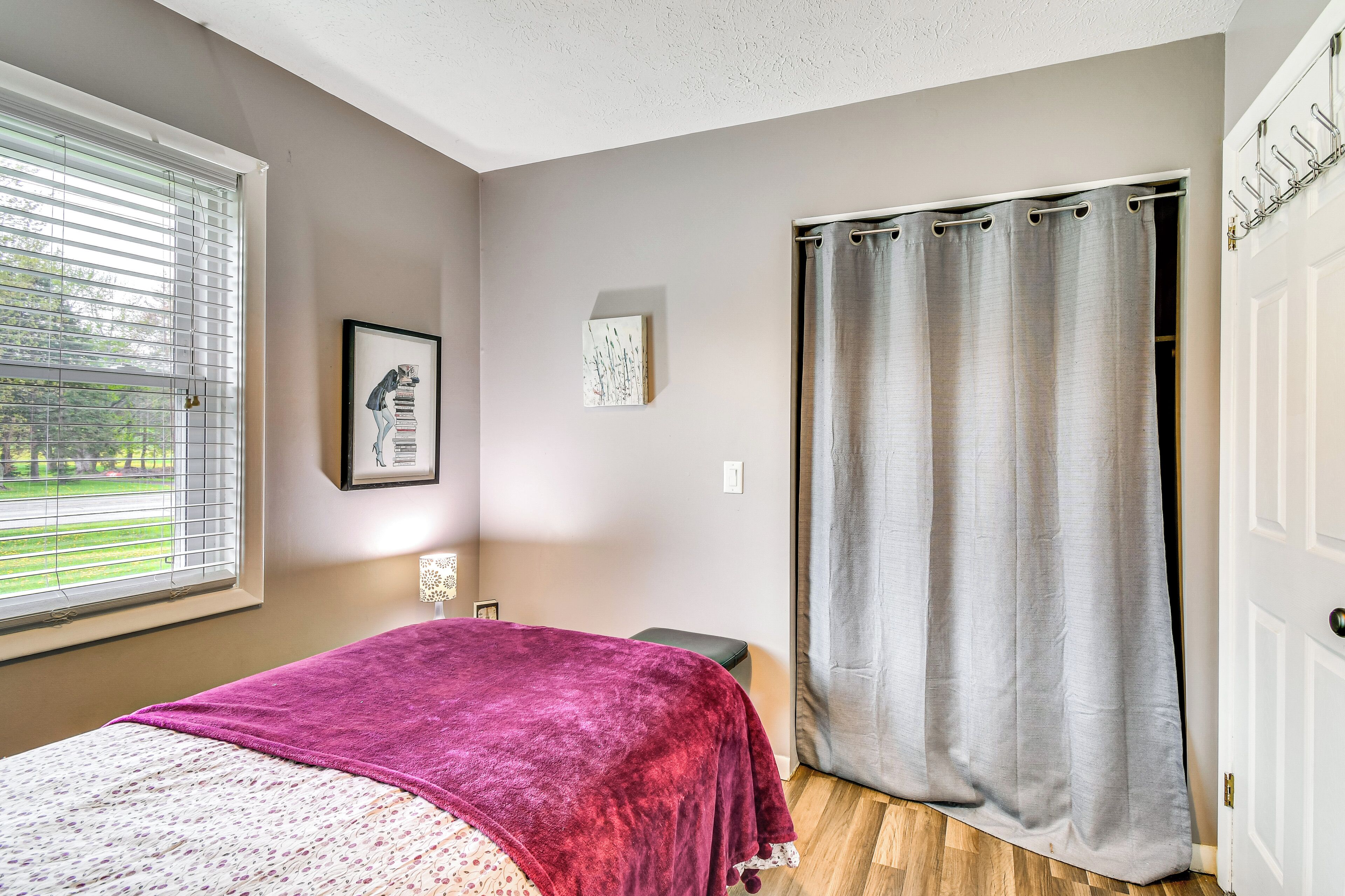 A bedroom with a bed, a window with blinds, a grey curtain, and a door with a coat rack.