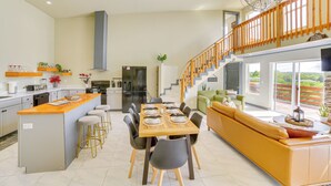 Una cocina amplia con una isla central, un comedor con mesa y sillas de madera, y una sala de estar luminosa con sofá amarillo y una escalera que sube a un nivel superior.
