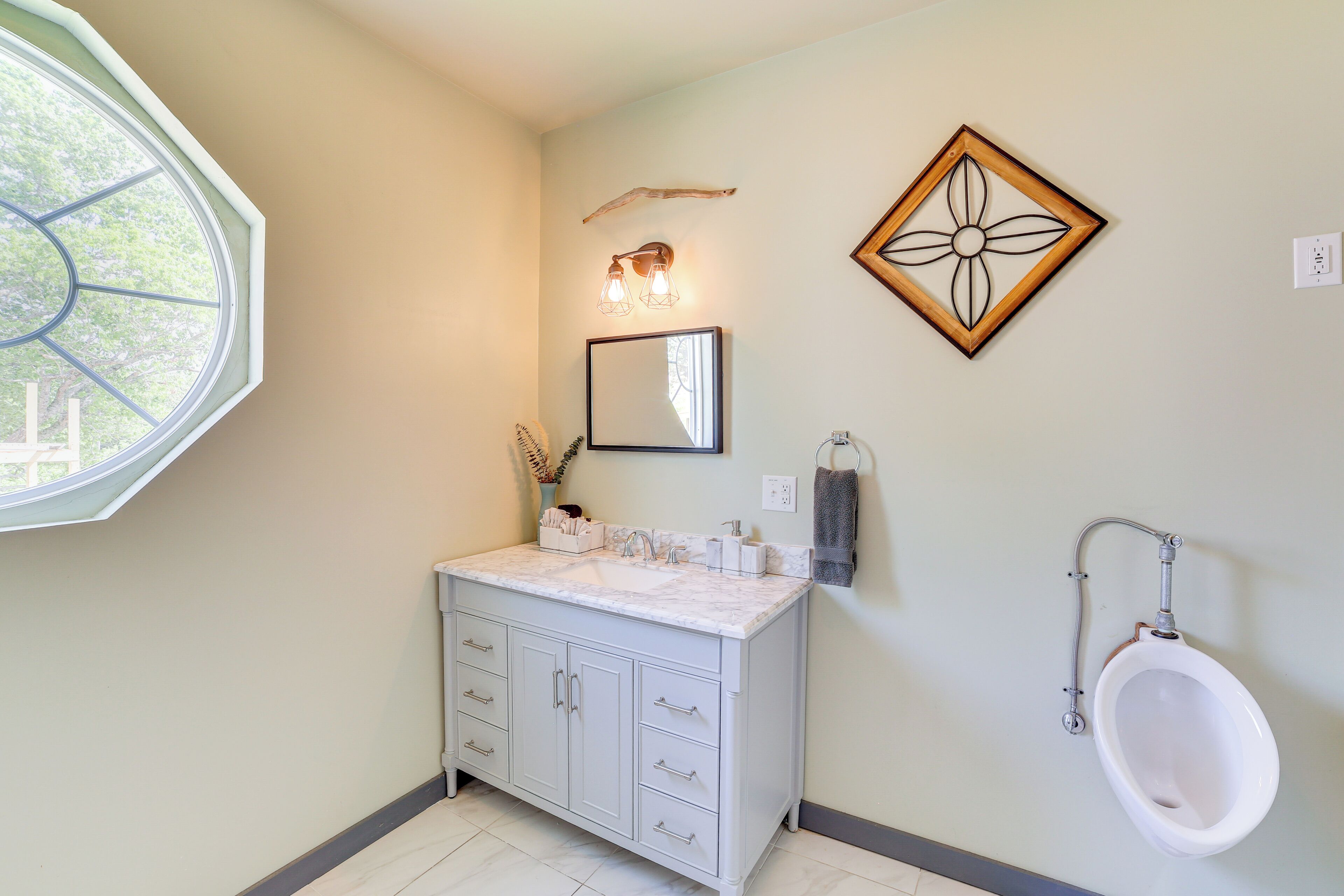 A bathroom with a white vanity, a mirror, a wall-mounted light fixture, a urinal, and a decorative wall piece.