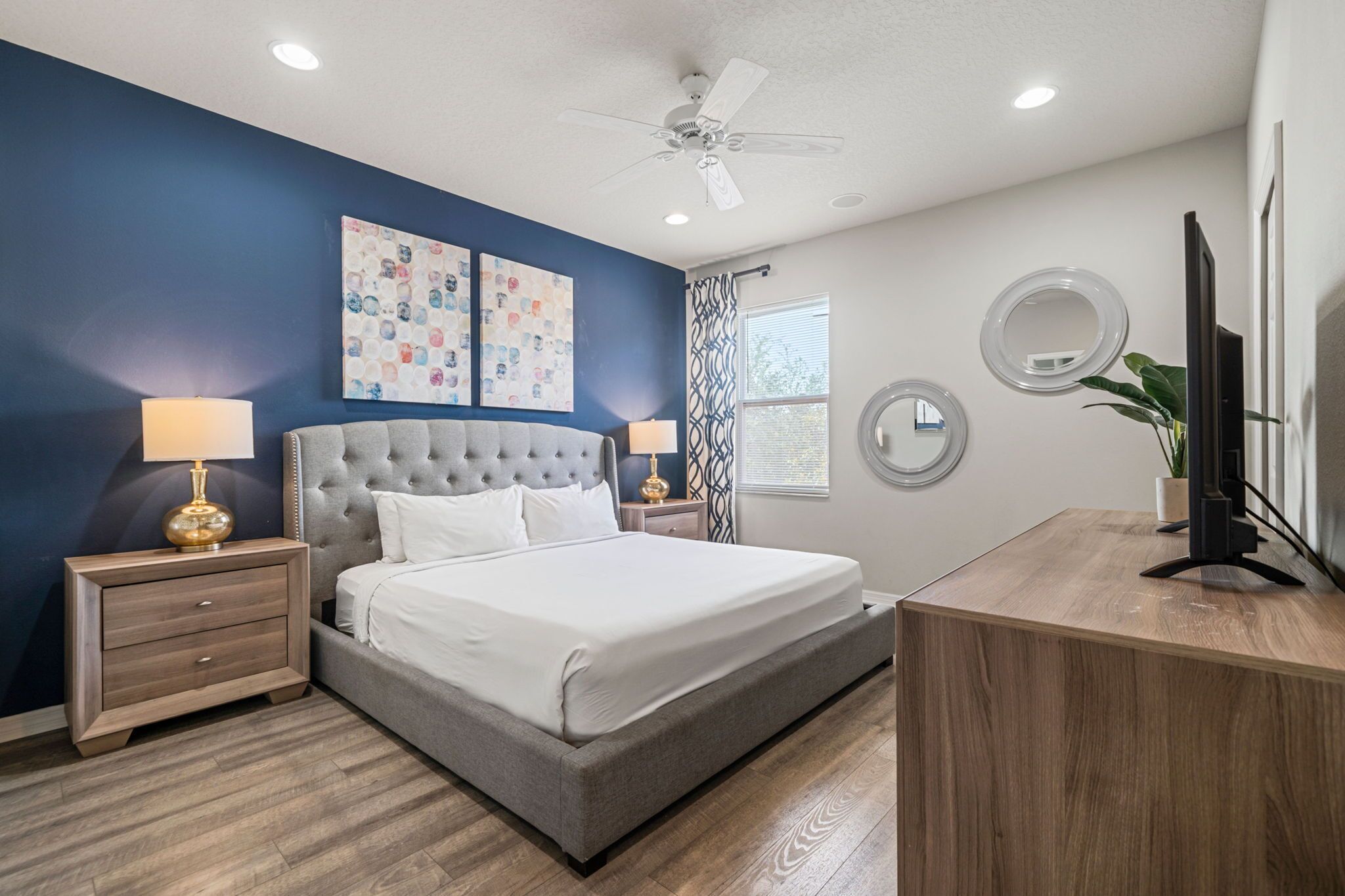 A bedroom with a large bed, two bedside tables with lamps, a ceiling fan, a TV, and artwork on the wall.