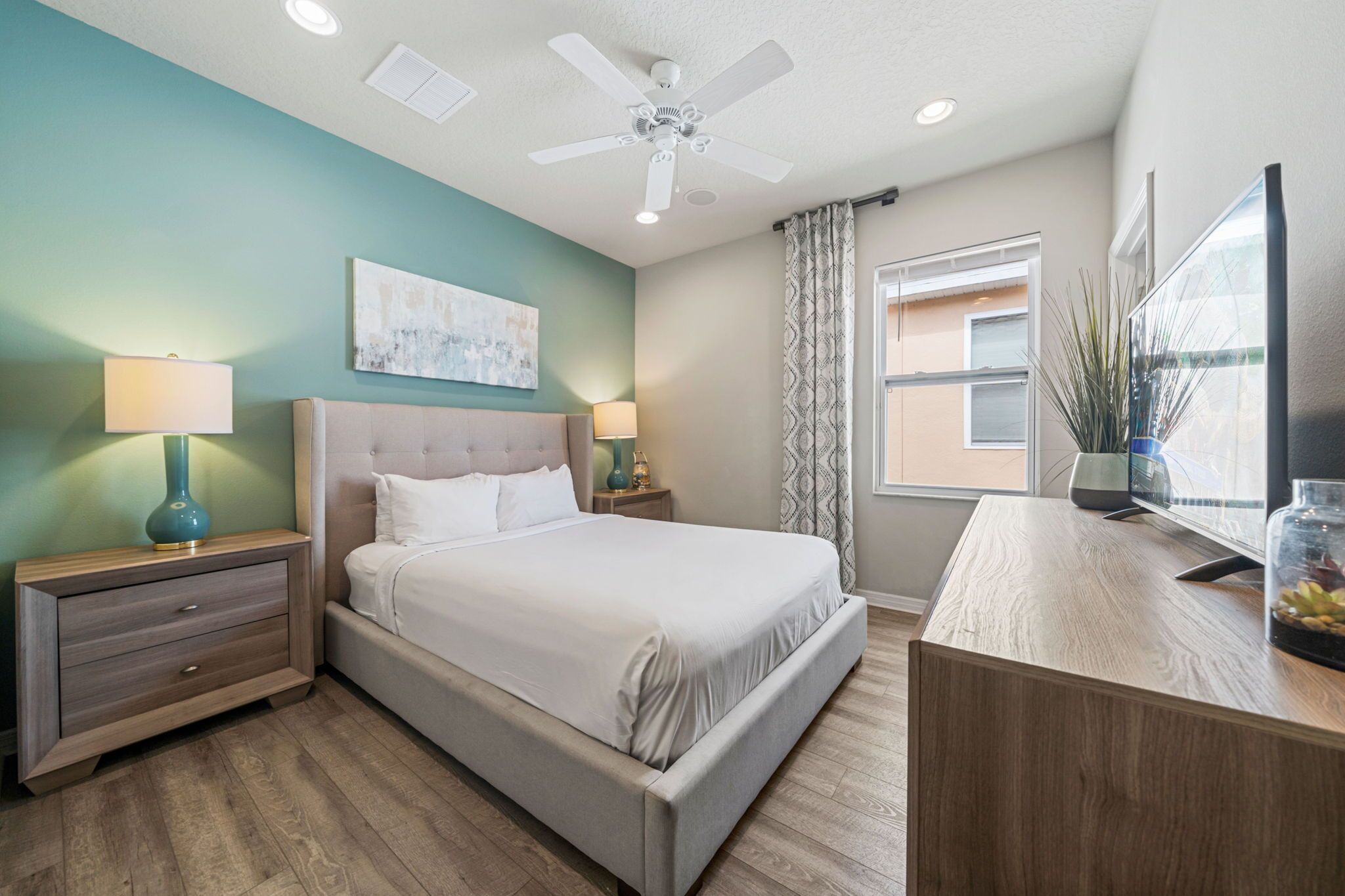 A bedroom with a bed, nightstands, a desk, a ceiling fan, a window with curtains, and a painting on the wall.