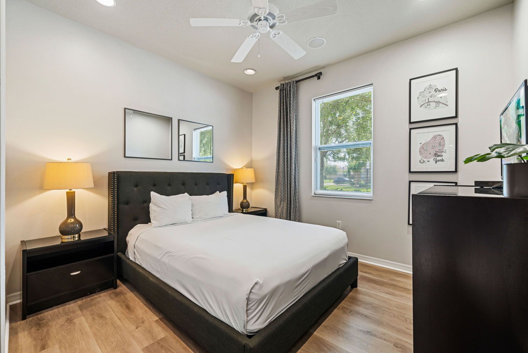 A modern bedroom with a large bed, two bedside tables with lamps, a ceiling fan, and framed pictures on the wall.