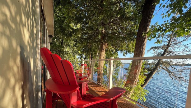 A wooden deck with red Adirondack chairs and a glass railing overlooking a body of water.
