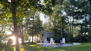 A cabin with a deck, surrounded by trees, and a fire pit in the yard.