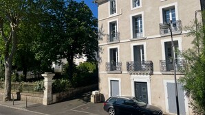Un immeuble de deux étages doté de balcons, une voiture garée devant et une rue bordée d’arbres.